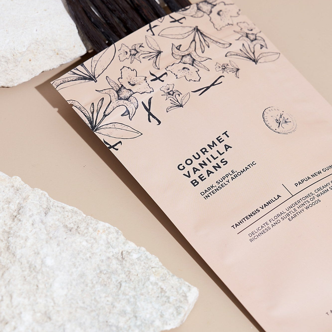 A pouch of Gourmet Vanilla Beans with vanilla beans sticking out, styled on a beige background with white textured stones. The pouch is a soft blush pink with black botanical illustrations and text. — Tahitian vanilla beans, Papua New Guinea